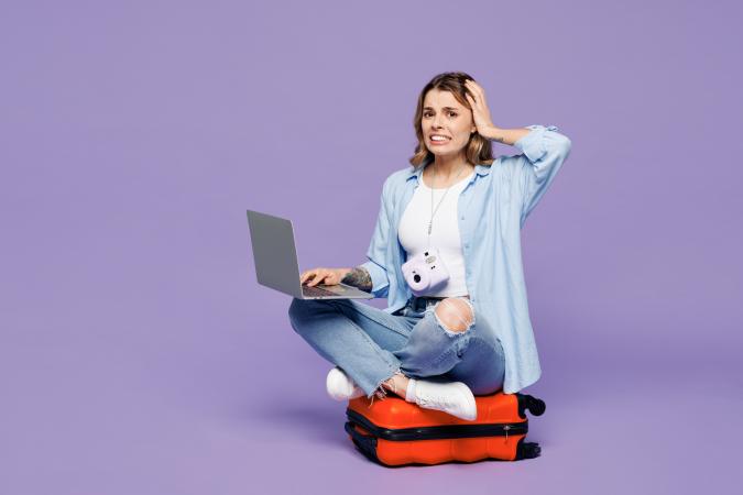 A worried young woman sitting cross-legged on a suitcase, holding a laptop on her lap, with a distressed expression and one hand on her head.