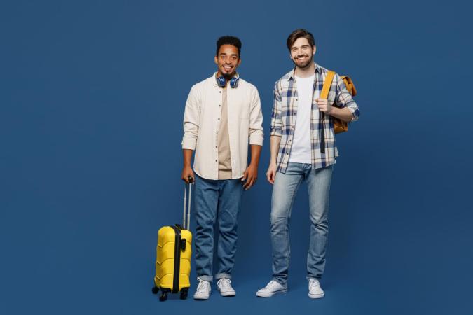 Two young men stand smiling, one holding a wheeled suitcase and the other carrying a backpack over one shoulder. Both appear ready for travel or an adventure