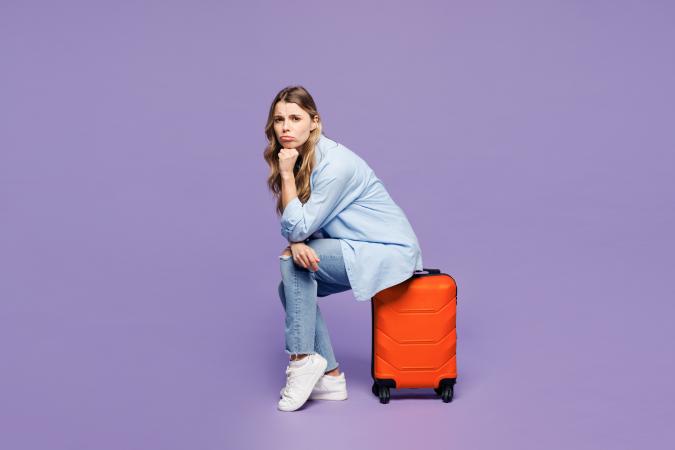 A young woman sitting on a suitcase, resting her chin on her hand with a thoughtful or slightly worried expression.