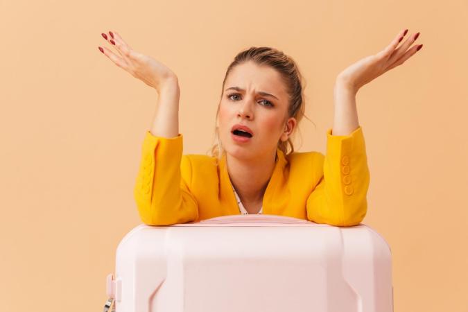 A frustrated woman leaning on a suitcase with her arms raised and mouth open, expressing confusion or annoyance.