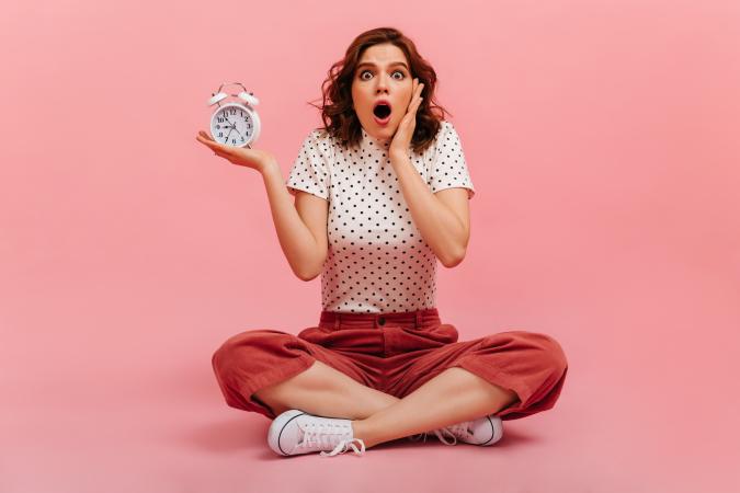 Een vrouw zit met gekruiste benen op de vloer, houdt een wekker in één hand vast en toont verrassing of schrik met een open mond, terwijl ze met haar andere hand haar wang raakt.