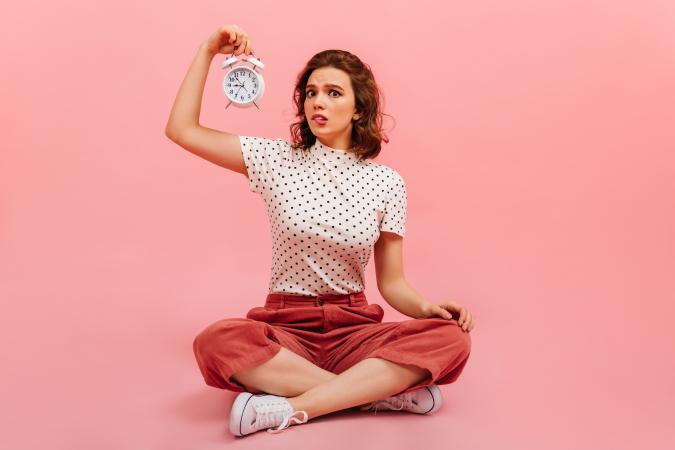 A young woman sitting cross-legged on the floor, holding an alarm clock up in one hand with a confused expression.