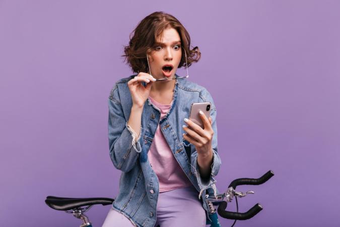 A young woman sitting on a bicycle, holding a smartphone in one hand, while removing her glasses with the other hand, showing a shocked or surprised expression.