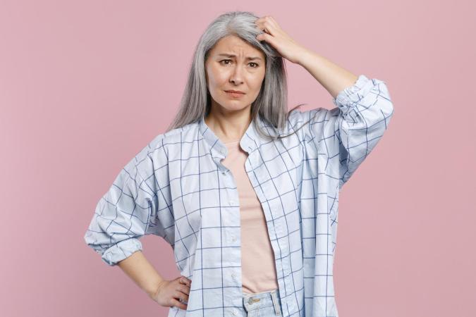 Een vrouw van middelbare leeftijd met lang haar staat met één hand op haar heup en de andere krabt aan haar hoofd. Ze kijkt verward of in de war.