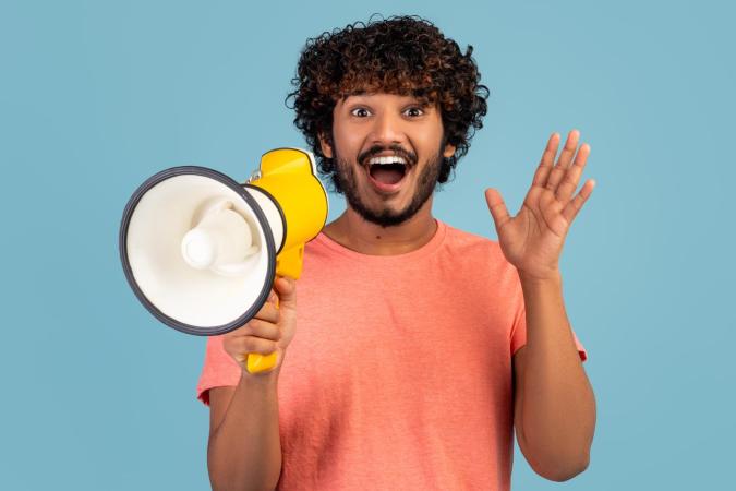 A young man with a curly hair and beard is holding a megaphone with one hand and waving with the other hand, looking excited and happy.