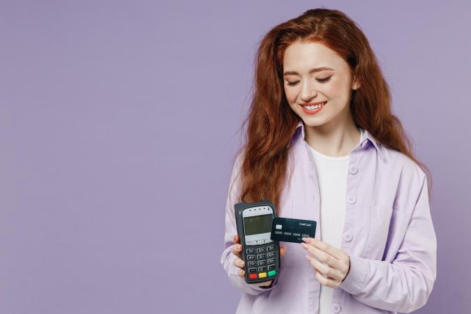 A woman smiling as she holds a payment terminal in one hand and a credit card in the other, preparing to make a transaction.