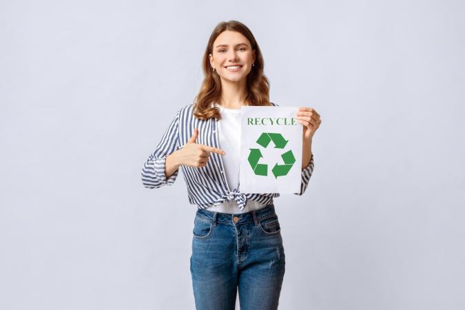A smiling woman is holding a paper with a recycling symbol and pointing at it, expressing enthusiasm and encouragement for recycling.