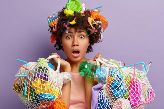 A surprised woman holding two mesh bags filled with various plastic items, while her hair is adorned with multiple small plastic objects, expressing shock or concern.