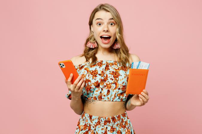 A young woman is smiling brightly while holding a smartphone in one hand and a passport with boarding passes in the other, expressing excitement and readiness for travel.