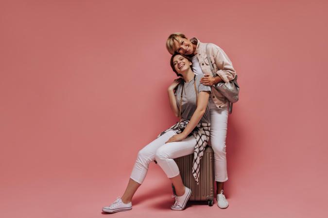 Twee vrouwen lachen blij en ontspannen. Eén zit op een koffer, de ander staat achter haar en leunt met haar hoofd tegen dat van de zittende vrouw. Ze omhelzen elkaar liefdevol.