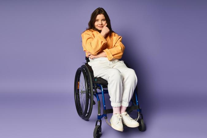 A young woman sitting in a wheelchair, resting her elbow on one armrest with her hand touching her chin, looking thoughtfully to the side with a slight smile.