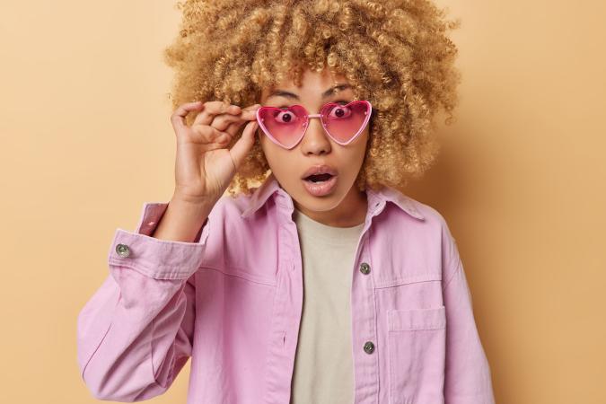 A young woman with curly hair and heart-shaped sunglasses looks surprised, holding one side of her glasses up as if to get a clearer view.