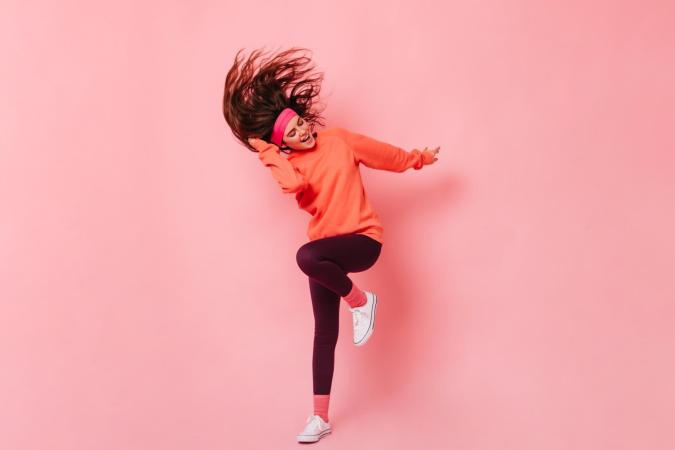 A young woman energetically dances with one leg raised, hair flipping, eyes closed, and a joyful expression on her face.