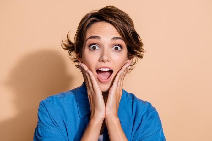 A woman with short hair holds her face with both hands, eyes wide open and mouth open, expressing surprise or amazement.