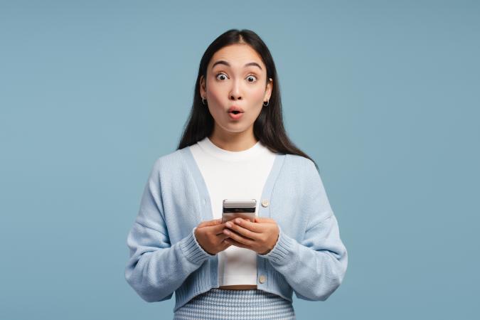 Vrouw kijkt verbaasd met grote ogen en ronde mond, houdt een telefoon vast met beide handen.