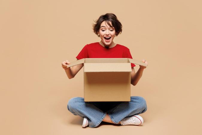 Een jonge vrouw zit met gekruiste benen op de vloer en houdt een open kartonnen doos voor zich, met een verraste en enthousiaste uitdrukking.