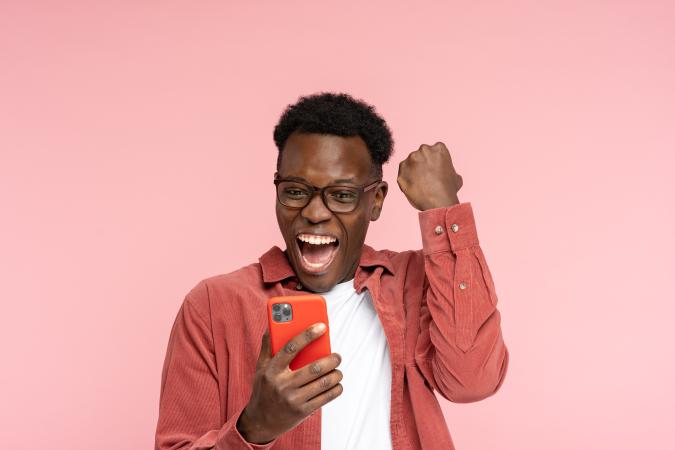 A joyful man wearing glasses looks at his phone with excitement, raising his fist in celebration.