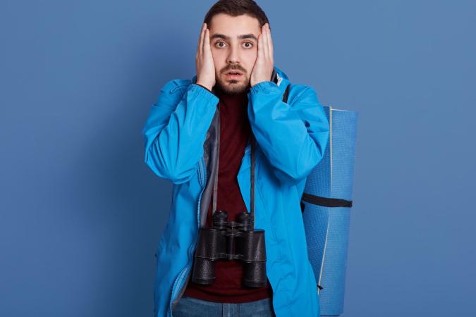 A man wearing outdoor gear looks shocked or worried, holding his face with both hands. He has binoculars hanging around his neck and a rolled-up mat strapped to his back.