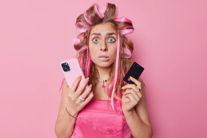 A young woman with hair curlers and smudged makeup holds a smartphone in one hand and a credit card in the other, looking confused and distressed.