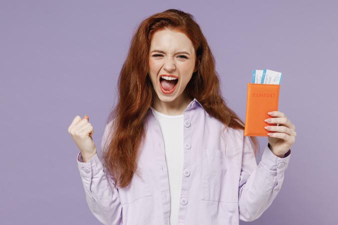 A young woman with long red hair shows excitement and triumph, holding a passport holder with flight tickets visible while making a fist with her other hand.