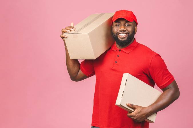 A smiling delivery man wearing a cap holds two cardboard boxes, one on his shoulder and the other in his hand, conveying readiness and friendliness.