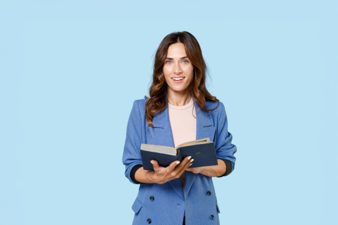 A smiling woman wearing a blazer holds an open book with both hands and looks directly ahead, appearing engaged and confident.