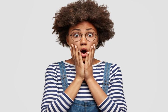 A woman wearing glasses holds her face with both hands, her mouth open in a surprised or shocked expression.