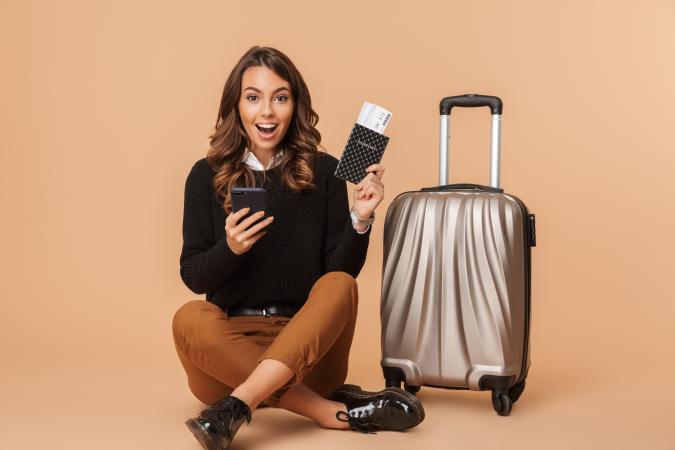 A young woman sitting cross-legged on the floor next to a suitcase, looking excited and surprised, holding a phone in one hand and a passport with a boarding pass in the other.