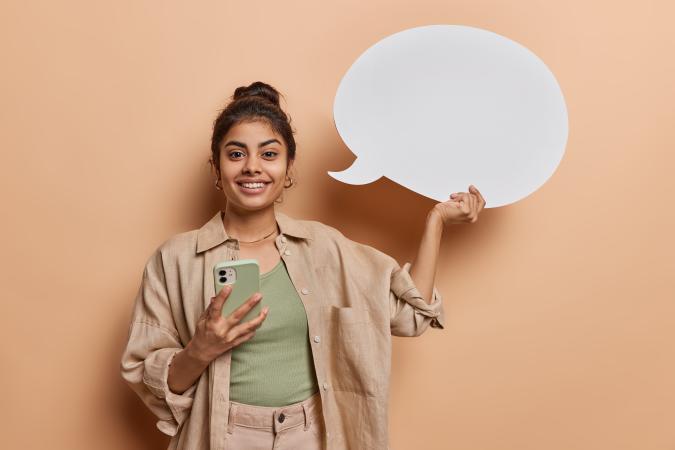 A young woman holding a phone smiles while holding up a speech bubble sign, expressing a positive and engaging demeanor.