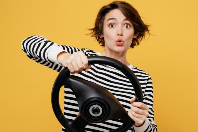 A woman holding a steering wheel with both hands, making a surprised or shocked face.
