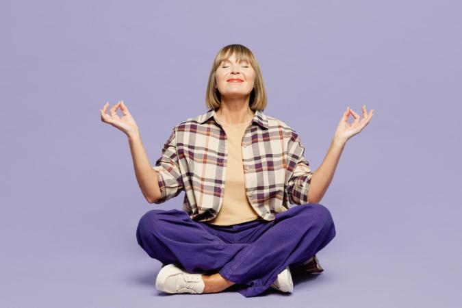 Een vrouw zit met gekruiste benen op de grond in een meditatieve houding. Haar ogen zijn gesloten en ze glimlacht rustig, terwijl ze haar handen ontspannen omhoog houdt met duim en wijsvinger tegen elkaar, alsof ze aan het mediteren is.