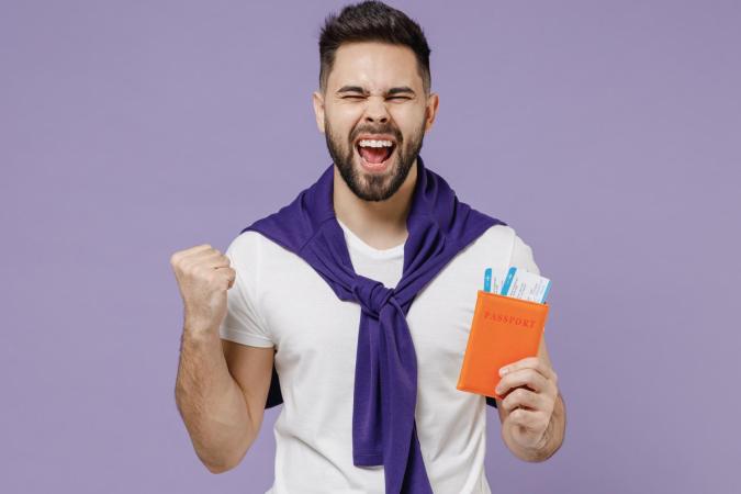 Een blije jonge man met een baard houdt een paspoorthoes vast met daarin twee instapkaarten. Met zijn andere hand maakt hij een feestelijk gebaar en zijn mond is open van blijdschap. 