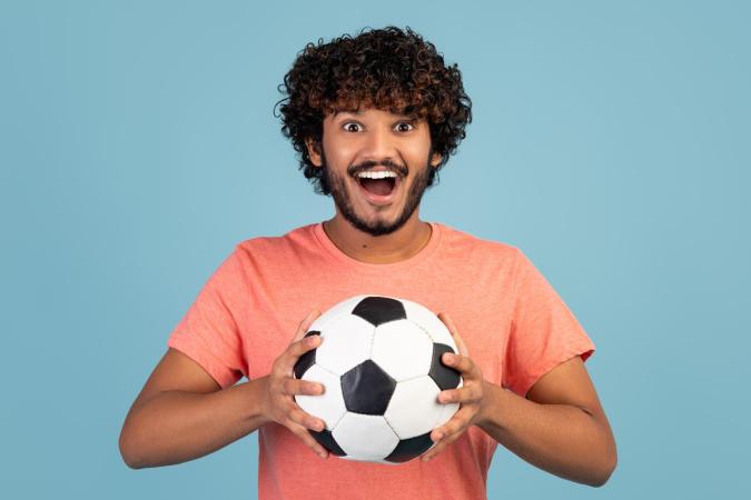 Een man houdt een voetbal met beide handen vast, kijkt enthousiast en glimlacht breed.