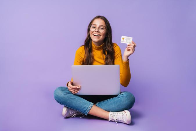 Meisje met een brede glimlach kijkt naar de camera, zittend met gekruiste benen. Ze houdt met haar rechterhand een creditcard omhoog en haar linkerhand rust op een laptop die op haar schoot staat.
