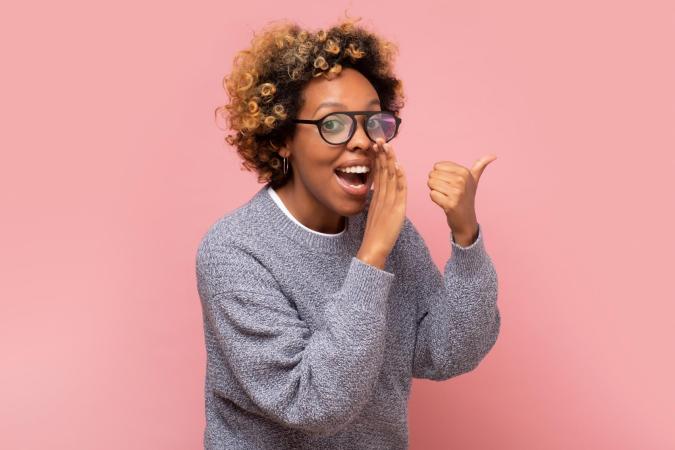 Vrouw met open mond alsof ze iets vertelt of roept, kijkt enthousiast. Ze houdt één hand voor haar mond en steekt met de andere hand haar duim omhoog.