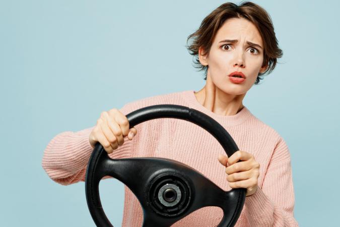 Een vrouw met een bezorgde of geschrokken blik houdt een stuur vast alsof ze aan het autorijden is. Ze kijkt recht voor zich uit met opgetrokken wenkbrauwen en een open mond.