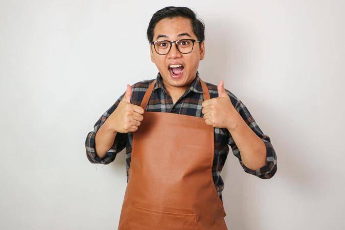 Een man met een bril en draagt een schort. Hij kijkt blij en enthousiast, met zijn mond open en geeft met beide handen een duim omhoog.