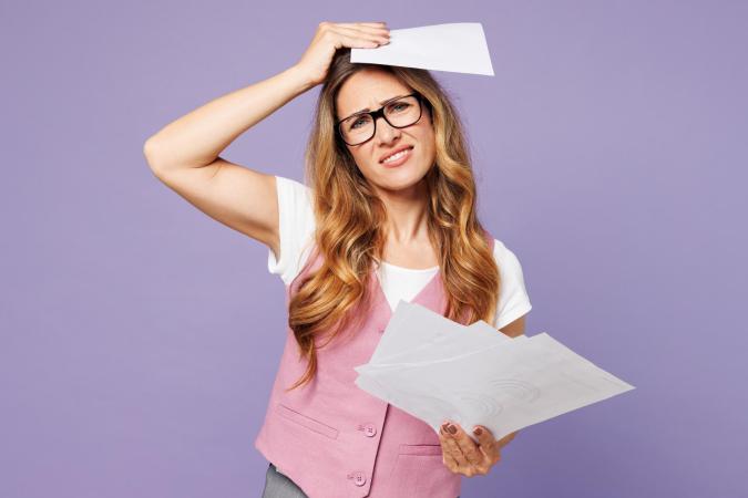 Vrouw kijkt gefrustreerd of bezorgd, trekt haar wenkbrauwen op en trekt haar mondhoek naar beneden. Ze houdt een stapel papieren in één hand en legt met de andere hand een vel papier tegen haar voorhoofd.