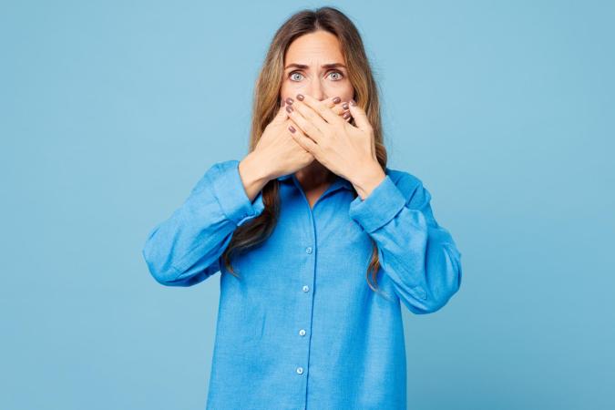 Vrouw kijkt geschrokken of bezorgd met grote ogen naar de camera. Ze bedekt haar mond met beide handen.
