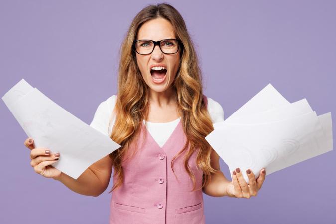 Vrouw kijkt boos en gefrustreerd met open mond. Ze houdt een stapel papieren in beide handen omhoog.