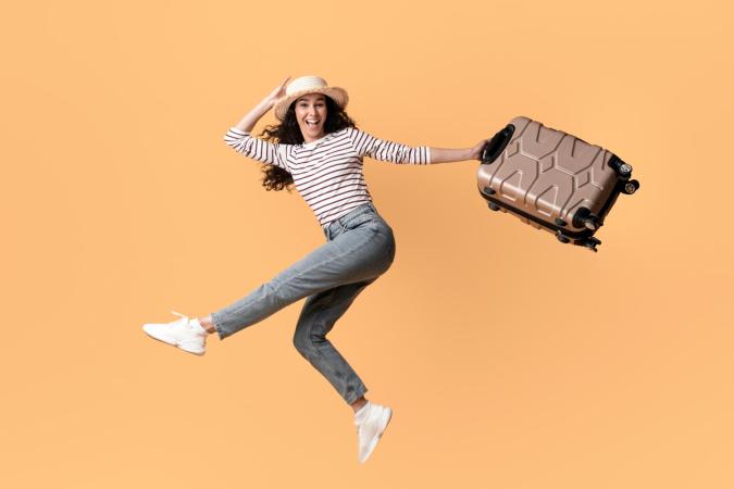 A joyful young woman holding a large tan rolling suitcase by its handle and smiling brightly