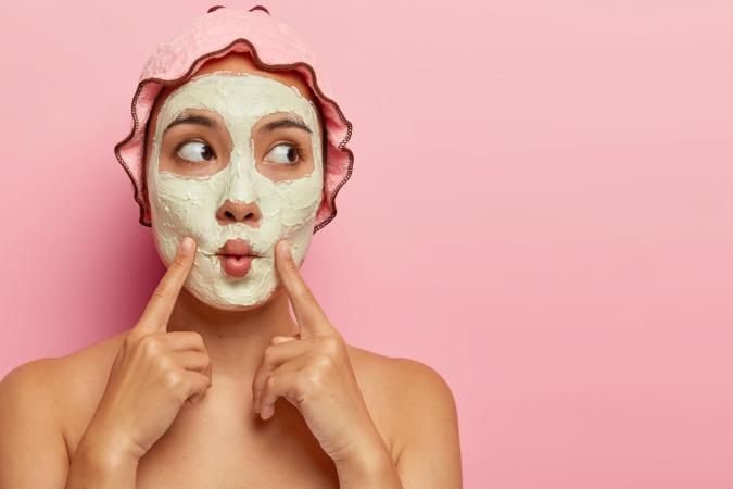 Vrouw met gezichtsmasker kijkt nieuwsgierig en speels opzij terwijl ze met haar wijsvingers haar gezicht aanraakt.