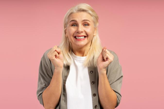Vrouw kijkt blij en enthousiast, met een brede glimlach en opgetrokken handen in een feestelijke of opgewonden houding.