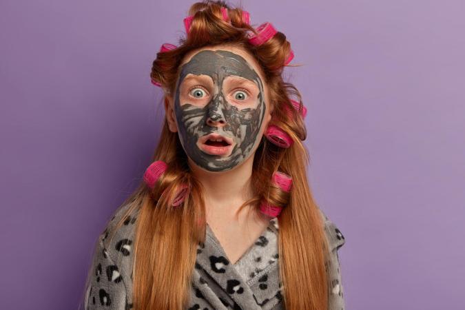 Vrouw kijkt verrast en verbaasd met haar mond iets open en ogen wijd open. Ze heeft haar hoofd bedekt met een kleimasker en draagt haar haar in krulspelden.