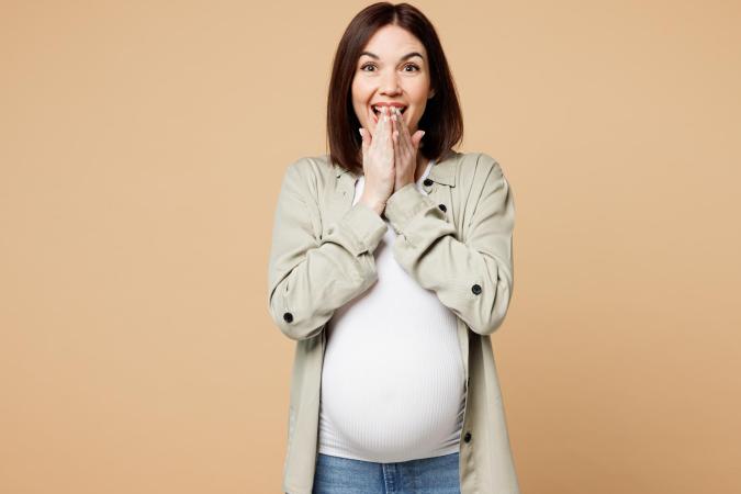 Zwangere vrouw kijkt blij en verrast, ze houdt haar handen voor haar mond en haar ogen zijn wijd open. Ze lijkt net een onverwacht leuk nieuws te hebben gehoord.