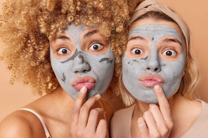 Twee vrouwen met gezichtsmaskers op, trekken een speelse, overdreven verbaasde gezichtsuitdrukking met grote ogen en licht vooruitgestoken lippen. Beiden raken met een vinger hun kin aan, alsof ze nadenken of iets grappigs overwegen.