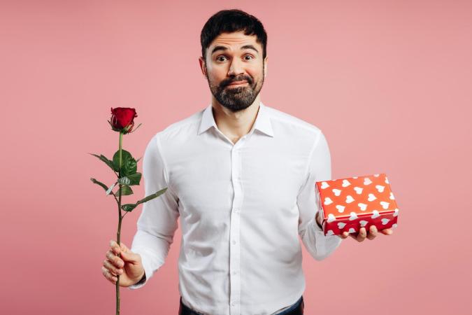 Een man met een lichte glimlach en opgetrokken wenkbrauwen kijkt direct naar de camera met een iets vragende of onzekere blik. Hij houdt in zijn rechterhand een enkele roos vast en in zijn linkerhand een cadeauverpakking met hartjesmotief, alsof hij iets aanbiedt of een keuze maakt.