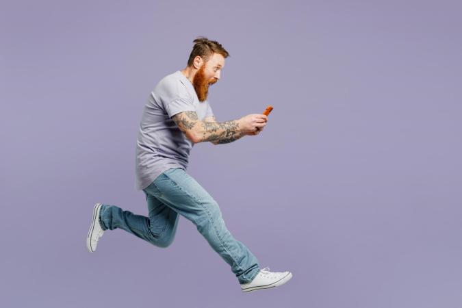 Een man die in de lucht lijkt te springen of te rennen, houdt zijn telefoon met beide handen vast en kijkt er met een grote open mond en verbaasde ogen naar, alsof hij iets onverwachts of spannends op het scherm ziet