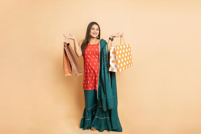 Een vrouw met een brede glimlach kijkt recht in de camera en houdt in elke hand meerdere tassen omhoog, alsof ze trots haar aankopen toont. Haar lichaamstaal straalt blijheid en tevredenheid uit.