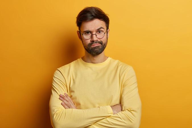 A man with beard, stands with his arms crossed. He has a slightly raised eyebrow and a skeptical or doubtful expression